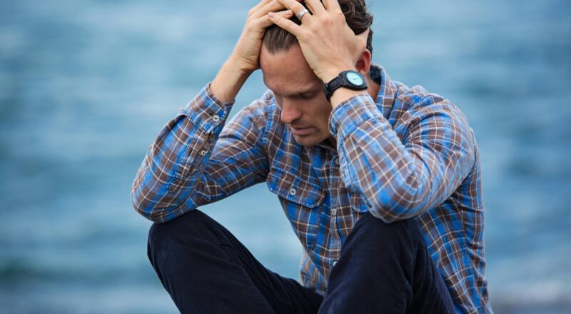 A man in a plaid shirt sits by the water looking distressed, symbolizing stress.